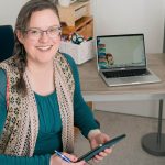 Nicole Lux-Ritche smiling and holding her phone at her desk in Luxcentric office